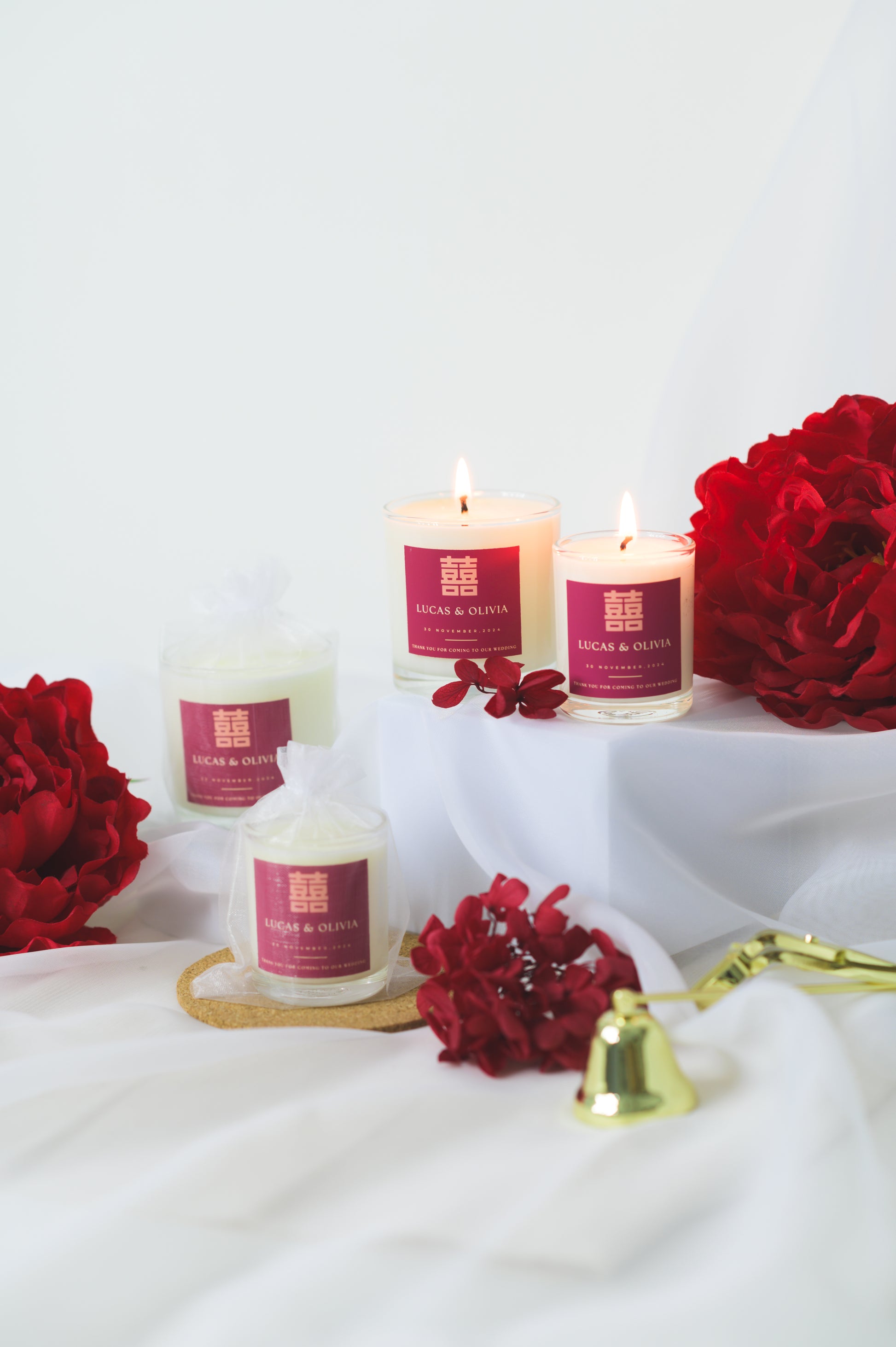 A lit custom wedding scented candle featuring a red 'Double Happiness' label with the names 'Lucas & Olivia,' wedding date, and a thank you message, placed beside red flowers as a wedding favor or gift.