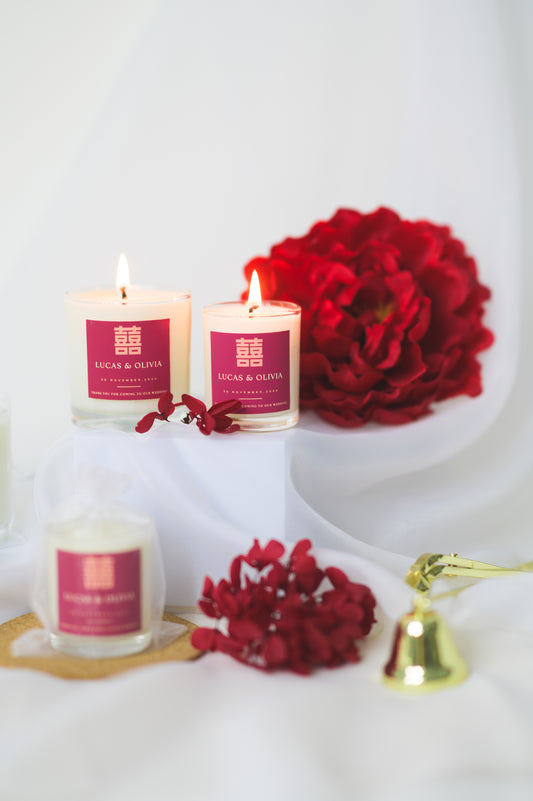 A lit custom wedding scented candle featuring a red 'Double Happiness' label with the names 'Lucas & Olivia,' wedding date, and a thank you message, placed beside red flowers as a wedding favor or gift.