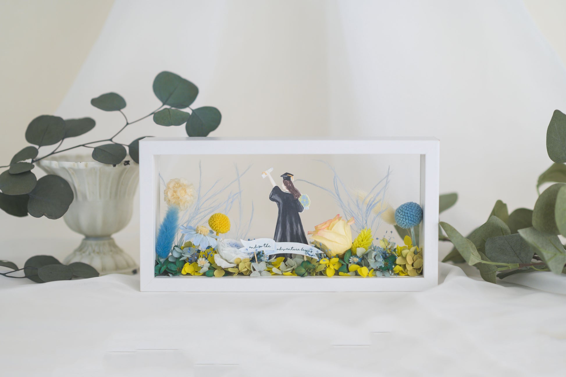 Graduation-themed preserved flower frame featuring a graduate figure holding a diploma, surrounded by vibrant yellow, blue, and white blooms.