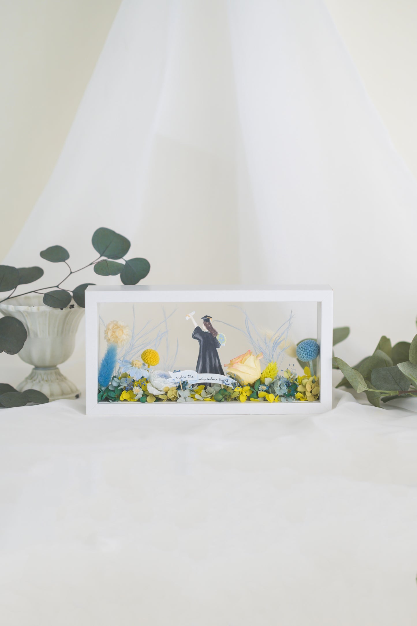Graduation-themed preserved flower frame featuring a graduate figure holding a diploma, surrounded by vibrant yellow, blue, and white blooms.