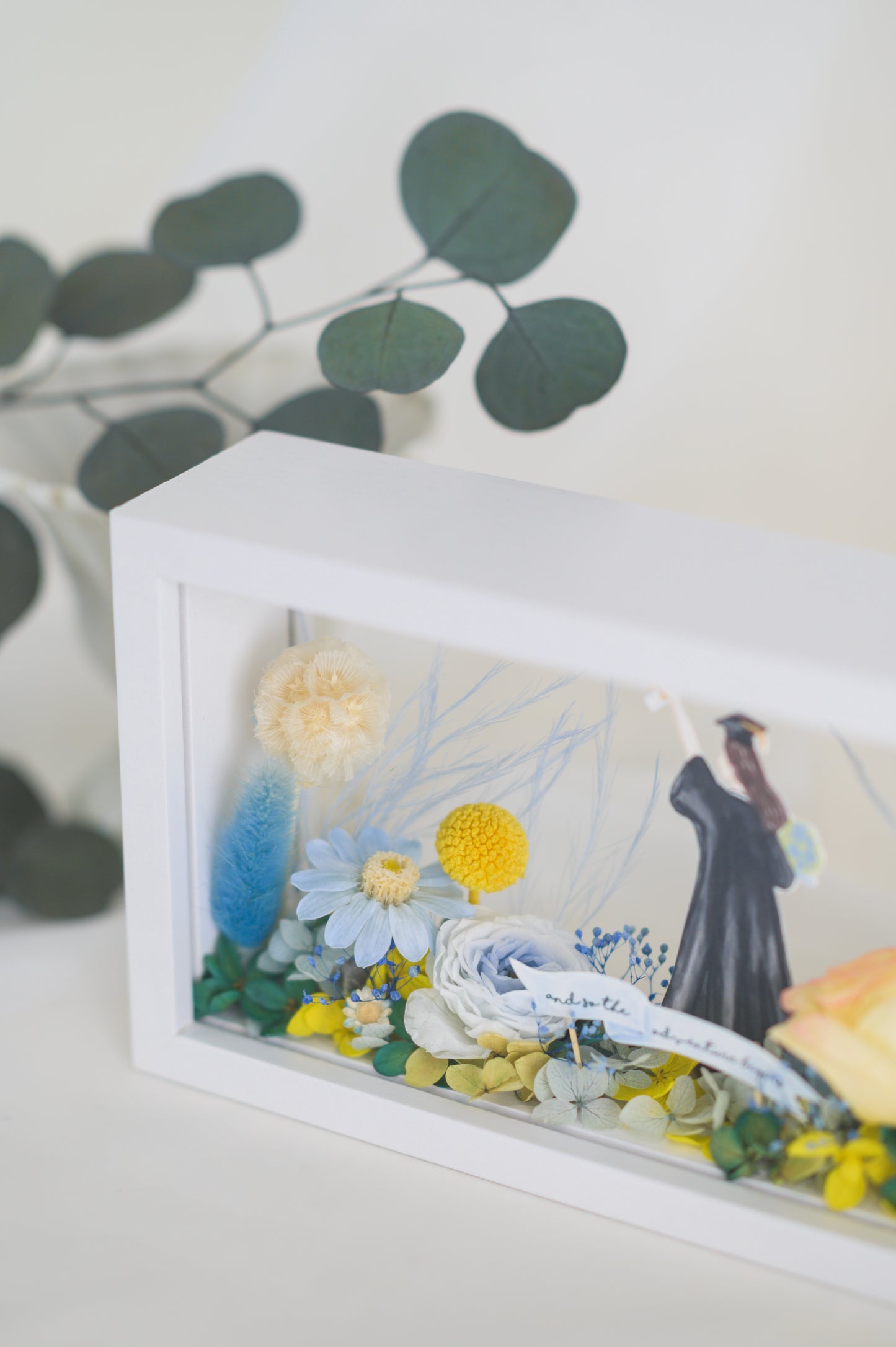 Graduation-themed preserved flower frame featuring a graduate figure holding a diploma, surrounded by vibrant yellow, blue, and white blooms.