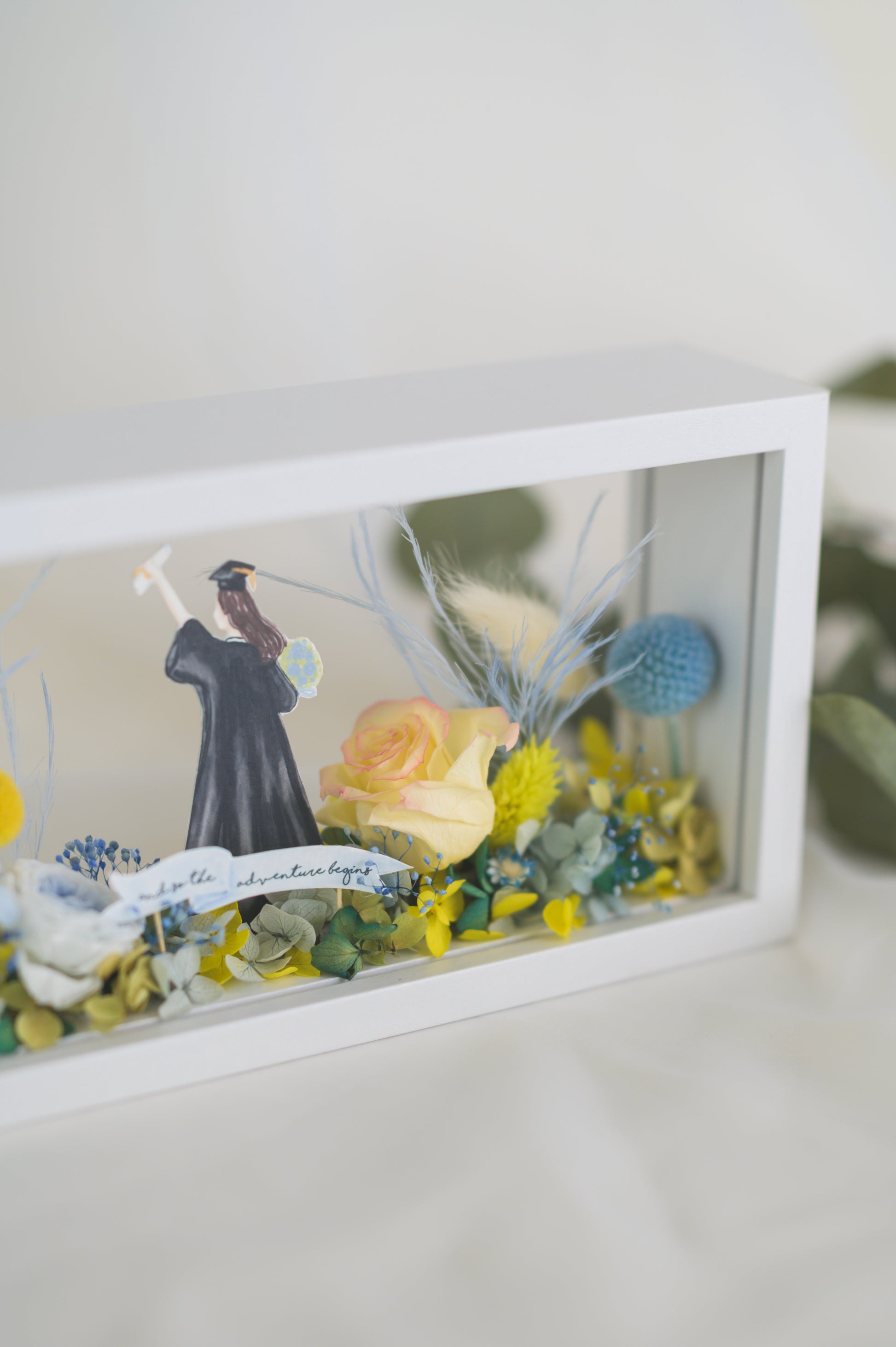 Graduation-themed preserved flower frame featuring a graduate figure holding a diploma, surrounded by vibrant yellow, blue, and white blooms.