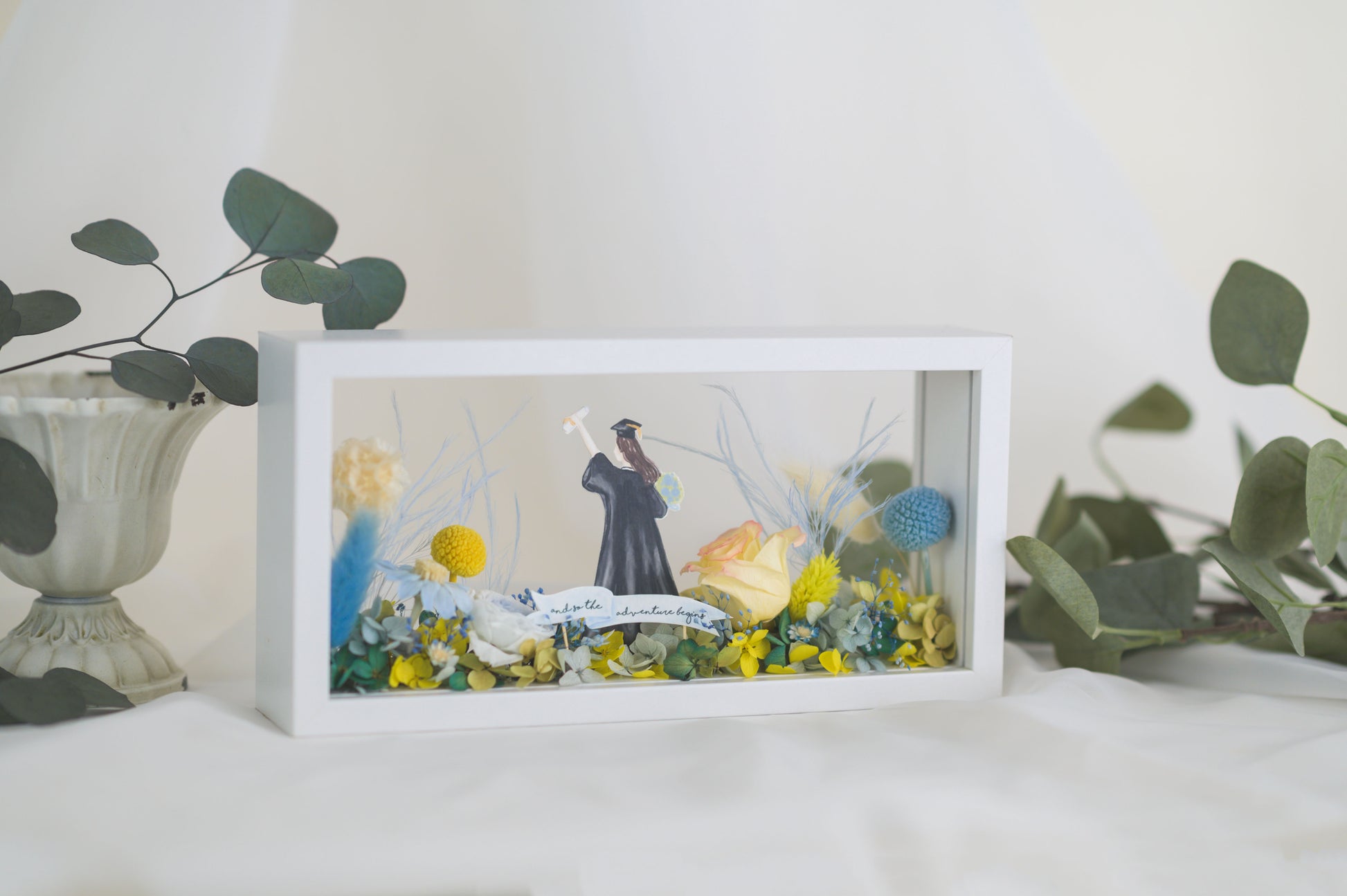 Graduation-themed preserved flower frame featuring a graduate figure holding a diploma, surrounded by vibrant yellow, blue, and white blooms.