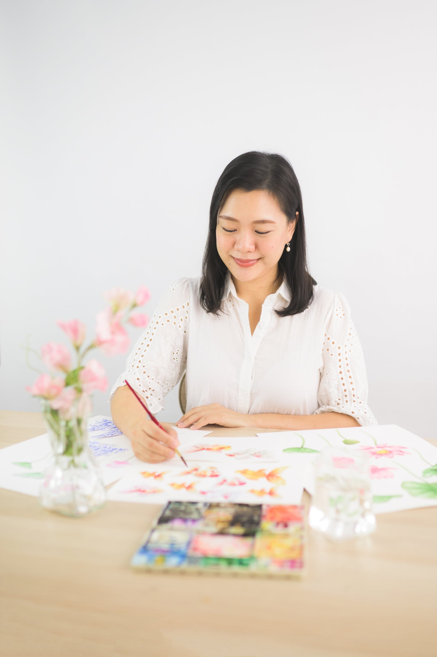 Artist Limzy working on a floral painting, seated at a table with watercolor supplies and pink flowers in a vase, wearing a white blouse and smiling as she creates her artwork.
