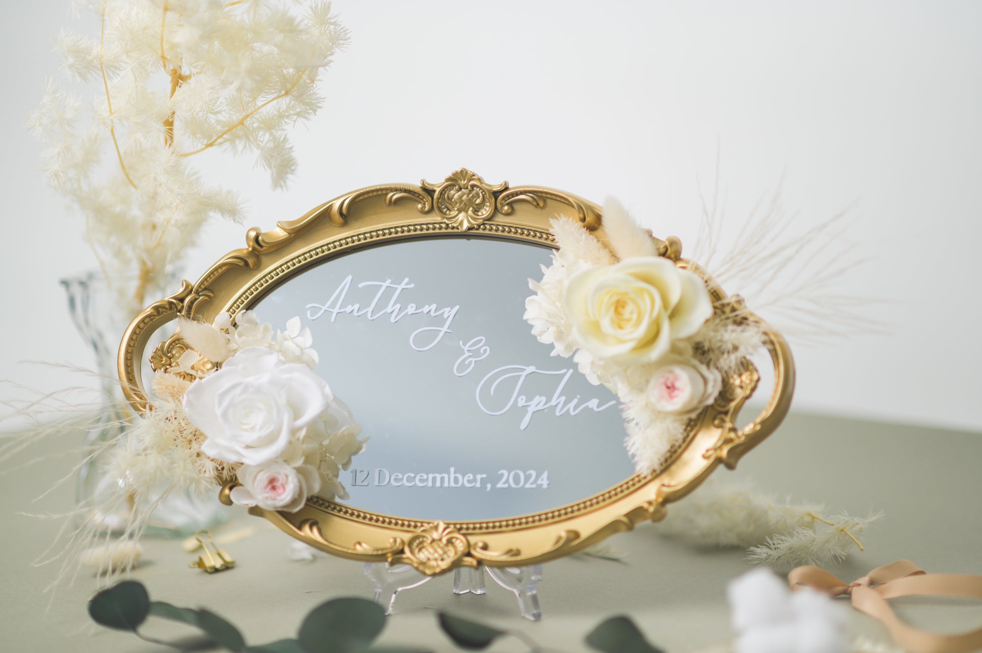 Ornate gold-framed wedding mirror featuring preserved white flowers, with customizable names Anthony & Sophia and wedding date, beautifully displayed on a reception table with floral accents.