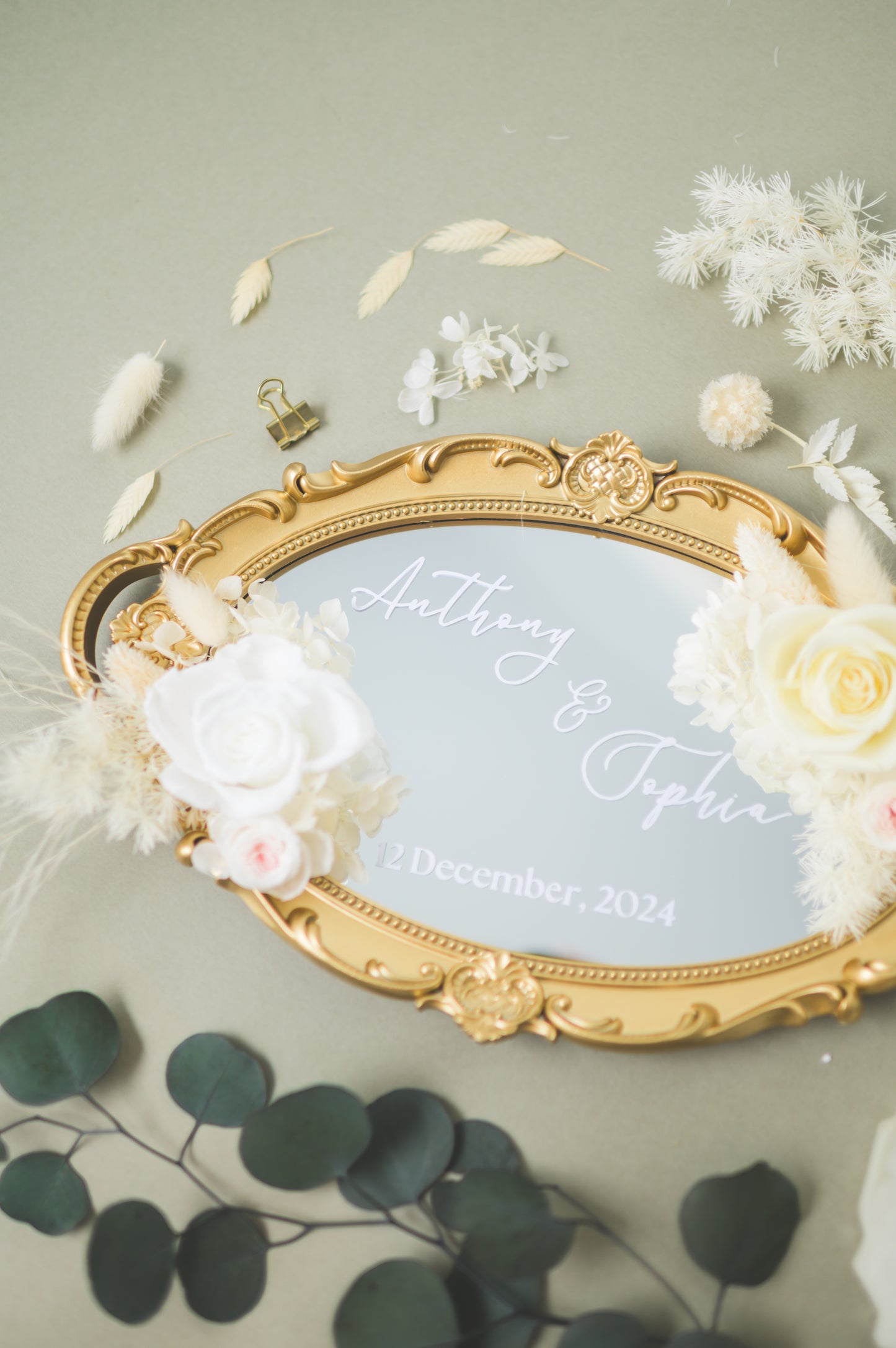 Ornate gold-framed wedding mirror featuring preserved white flowers, with customizable names Anthony & Sophia and wedding date, beautifully displayed on a reception table with floral accents.