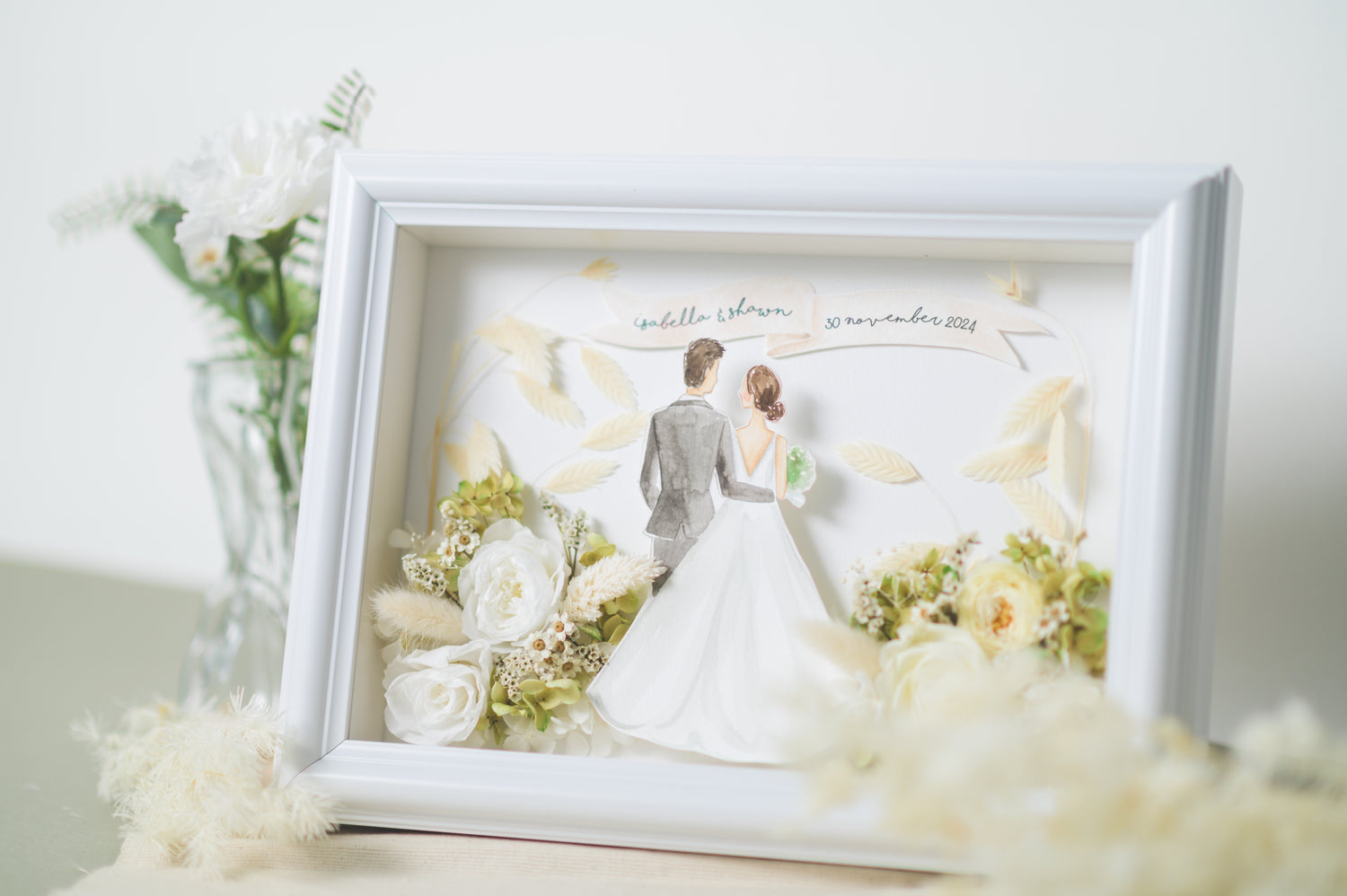 Personalized wedding frame featuring a bride and groom illustration with preserved white and green flowers, custom names and date, framed in white.
