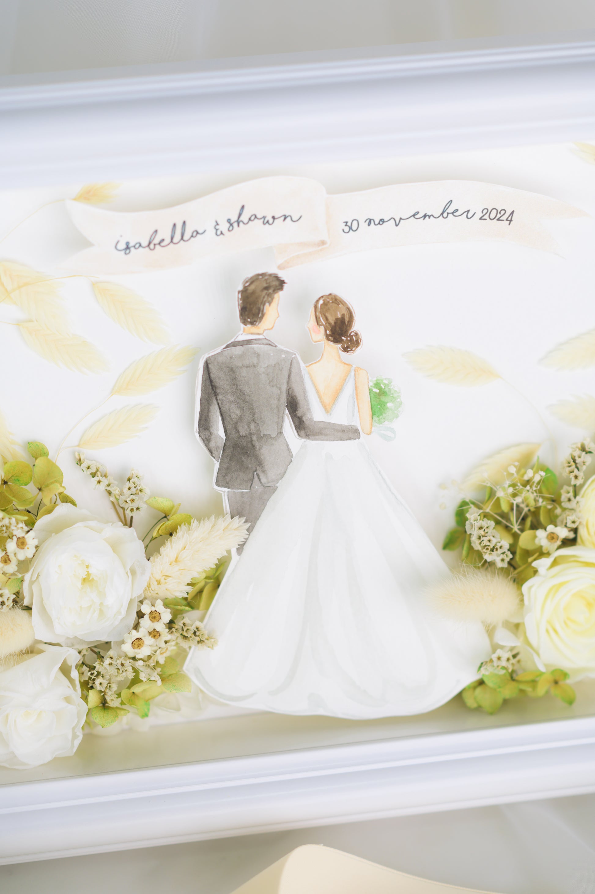 Personalized wedding frame featuring a bride and groom illustration with preserved white and green flowers, custom names and date, framed in white.