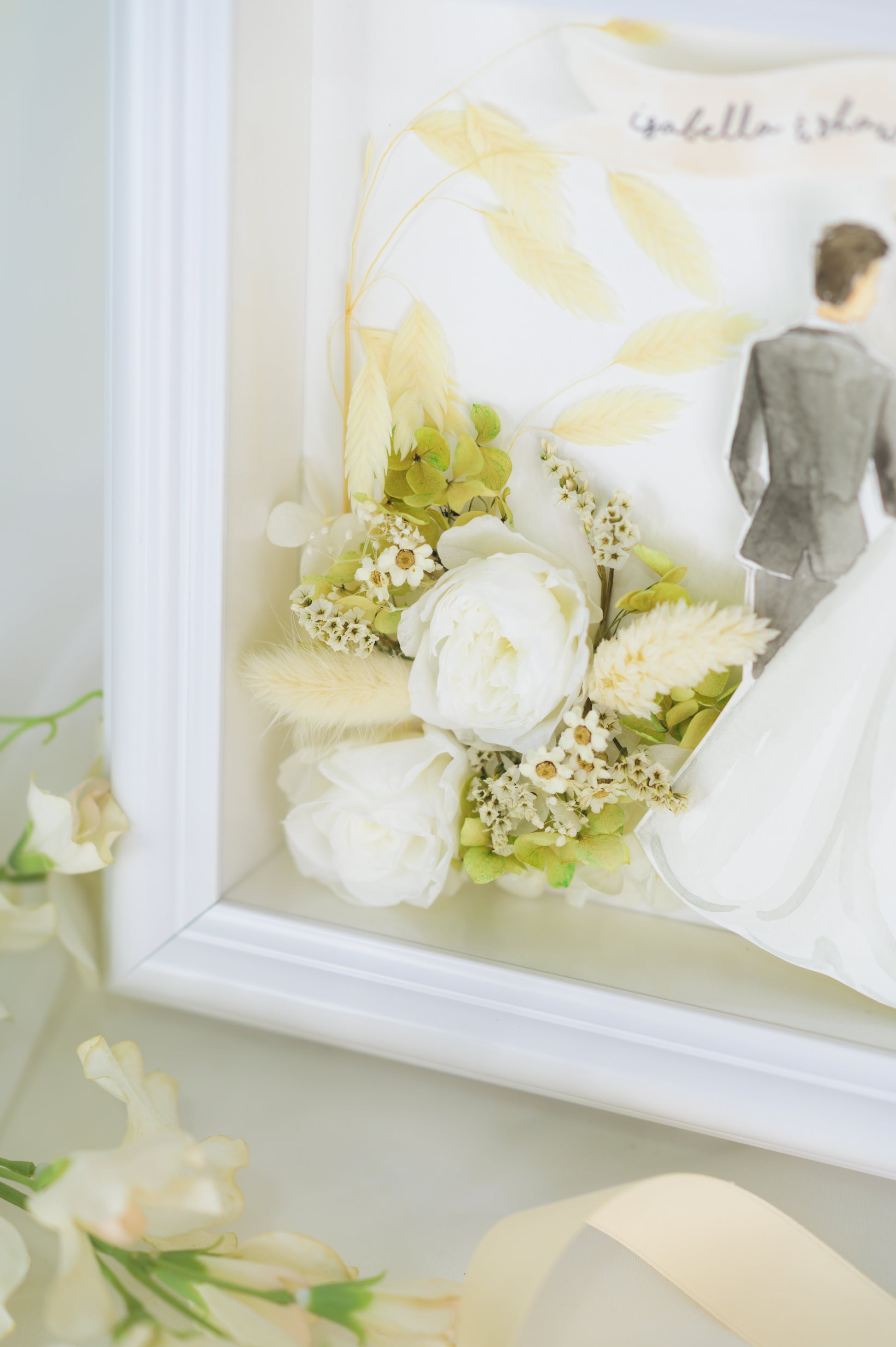 Personalized wedding frame featuring a bride and groom illustration with preserved white and green flowers, custom names and date, framed in white.