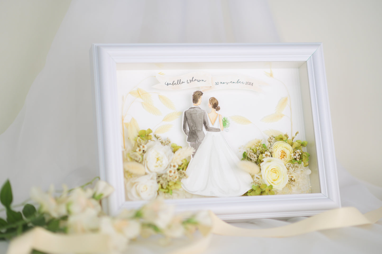 Personalized wedding frame featuring a bride and groom illustration with preserved white and green flowers, custom names and date, framed in white.
