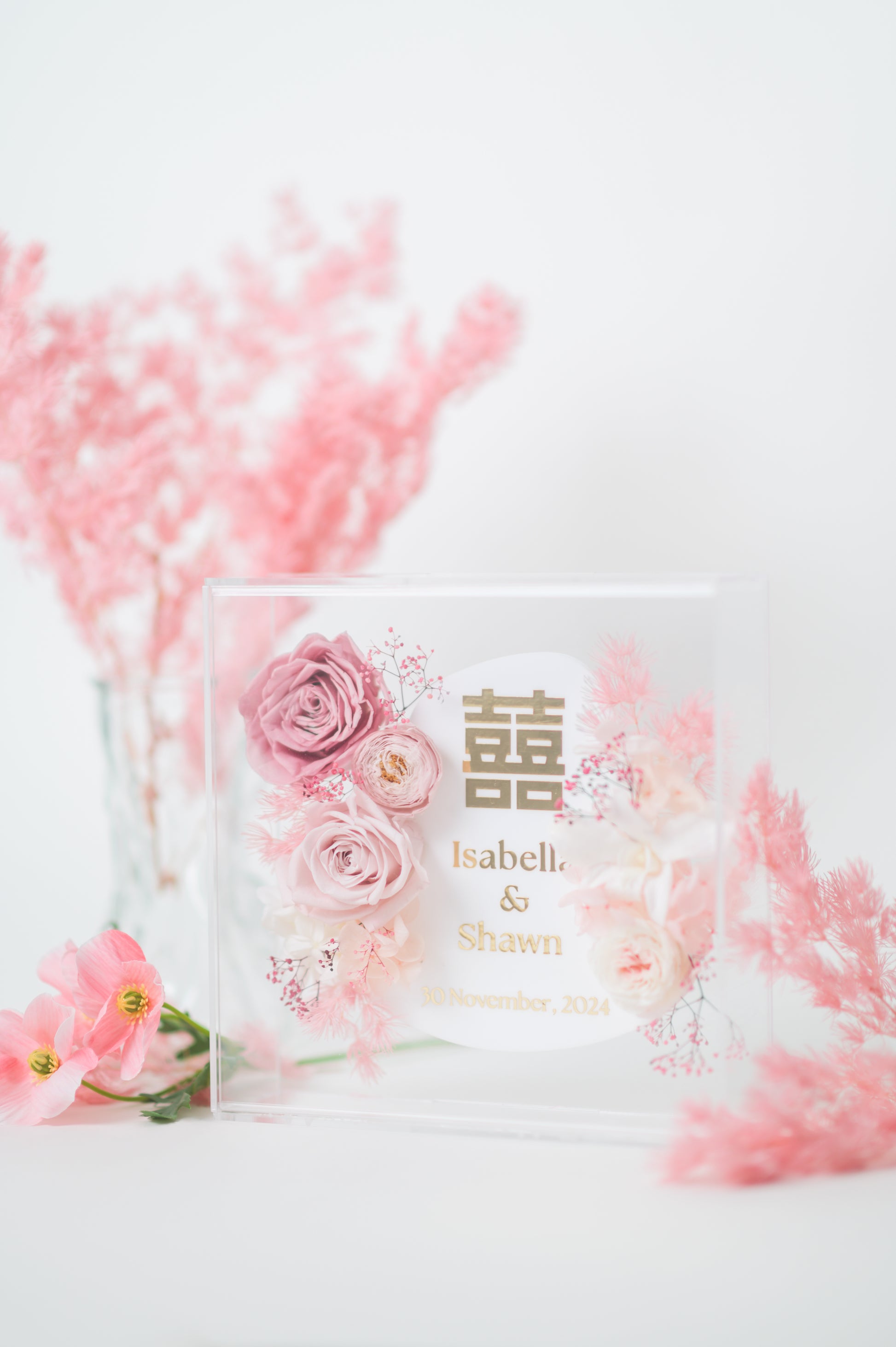 Personalized wedding frame with preserved pink flowers, featuring custom names Isabella & Shawn and a wedding date, with elegant floral design in a clear acrylic display.