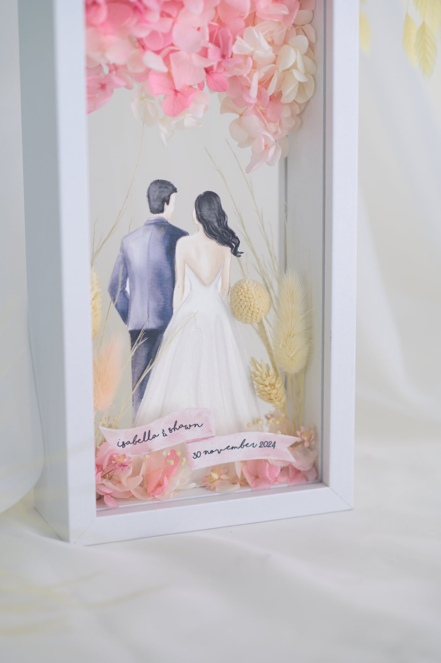 A custom wedding keepsake frame featuring a couple illustration in wedding attire, surrounded by preserved flowers in soft pinks and yellows. The frame has a front and back view, offering a beautiful display of the artwork. Customizable with names and date for a personal touch.