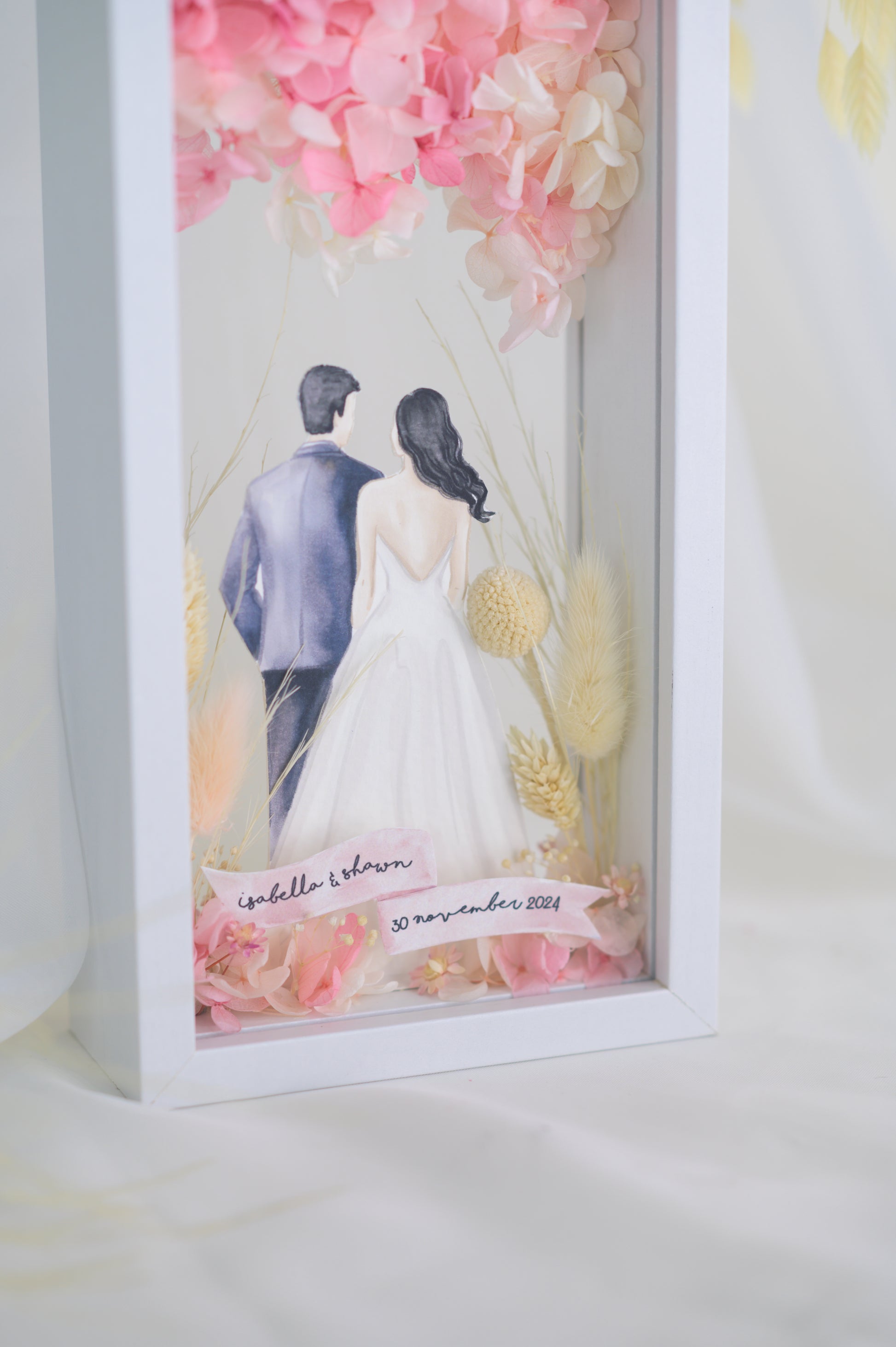 A custom wedding keepsake frame featuring a couple illustration in wedding attire, surrounded by preserved flowers in soft pinks and yellows. The frame has a front and back view, offering a beautiful display of the artwork. Customizable with names and date for a personal touch.
