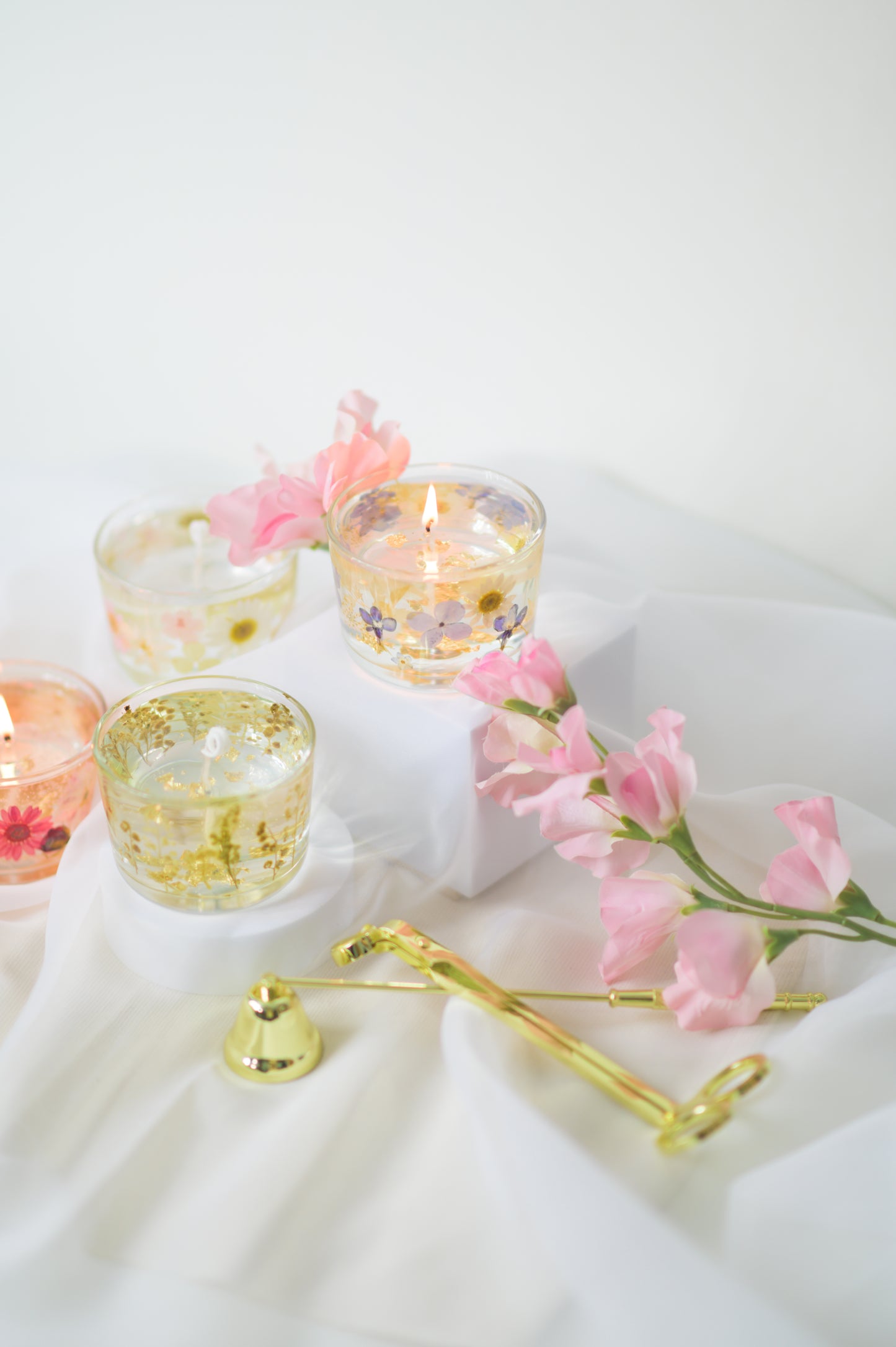 Handcrafted floral gel wax candles with embedded dried flowers and gold foil accents, burning gently on a soft fabric surface with a candle snuffer nearby.