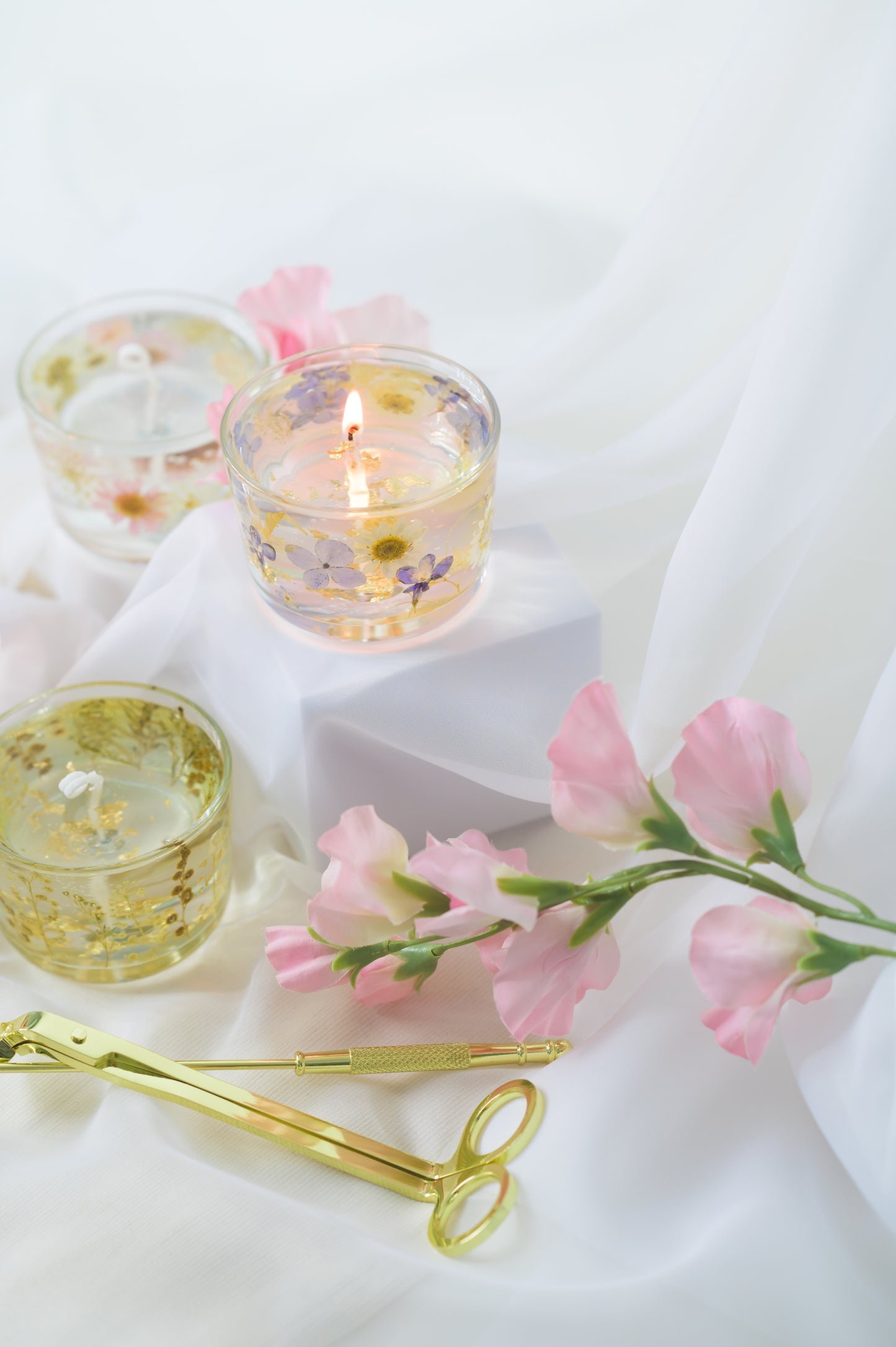 Handcrafted floral gel wax candles with embedded dried flowers and gold foil accents, burning gently on a soft fabric surface with a candle snuffer nearby.