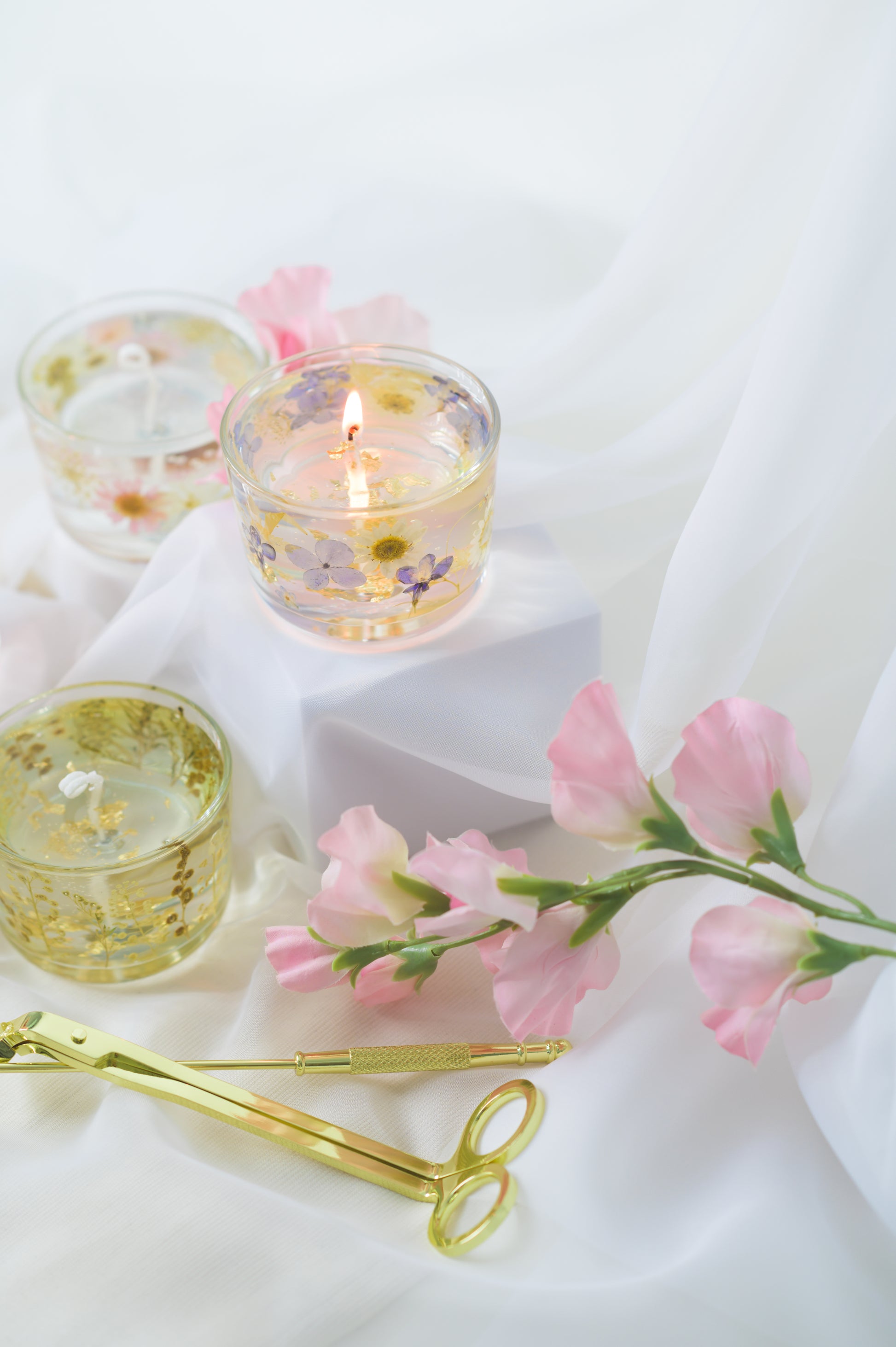 Handcrafted floral gel wax candles with embedded dried flowers and gold foil accents, burning gently on a soft fabric surface with a candle snuffer nearby.
