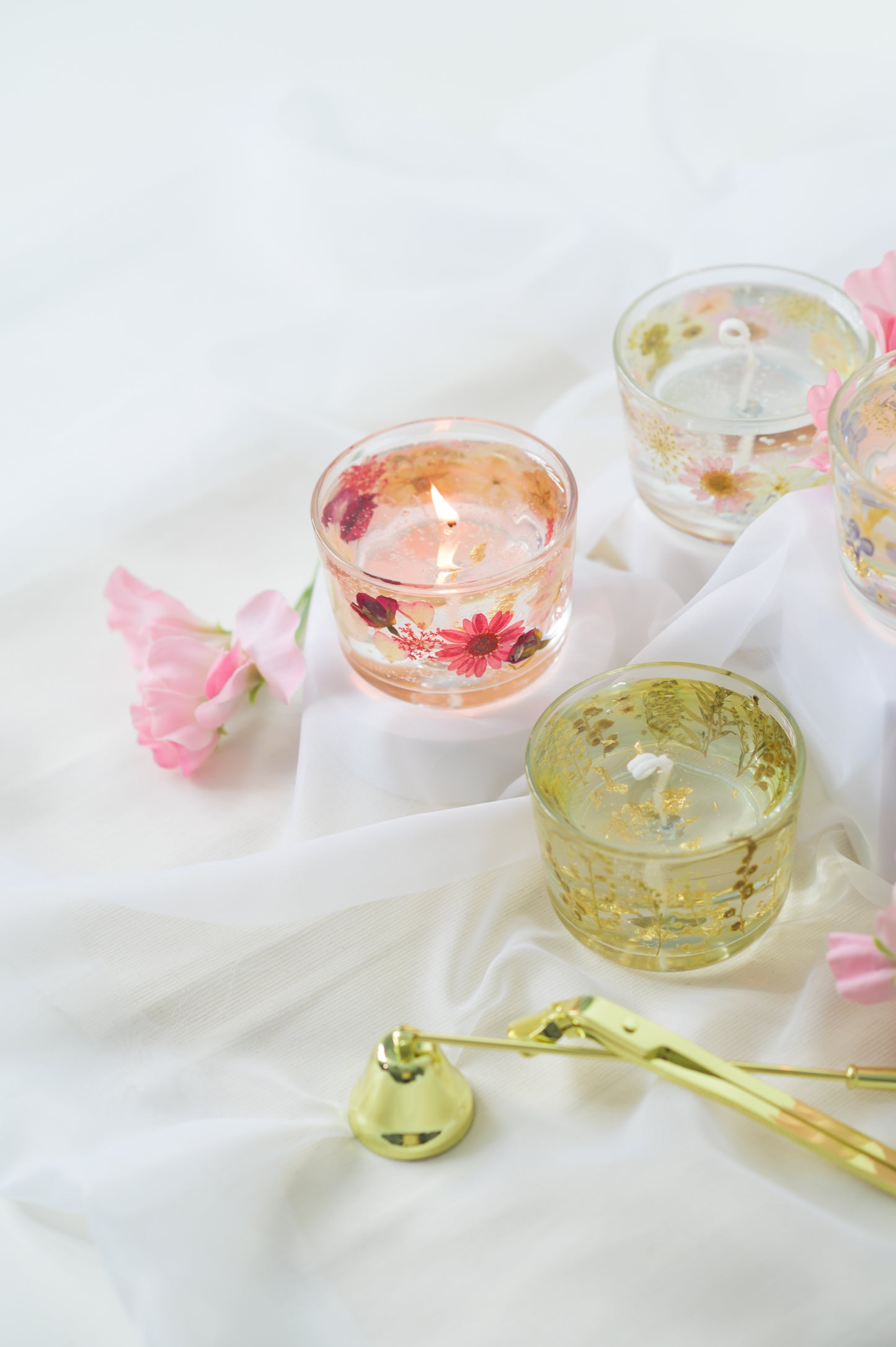 Handcrafted floral gel wax candles with embedded dried flowers and gold foil accents, burning gently on a soft fabric surface with a candle snuffer nearby.